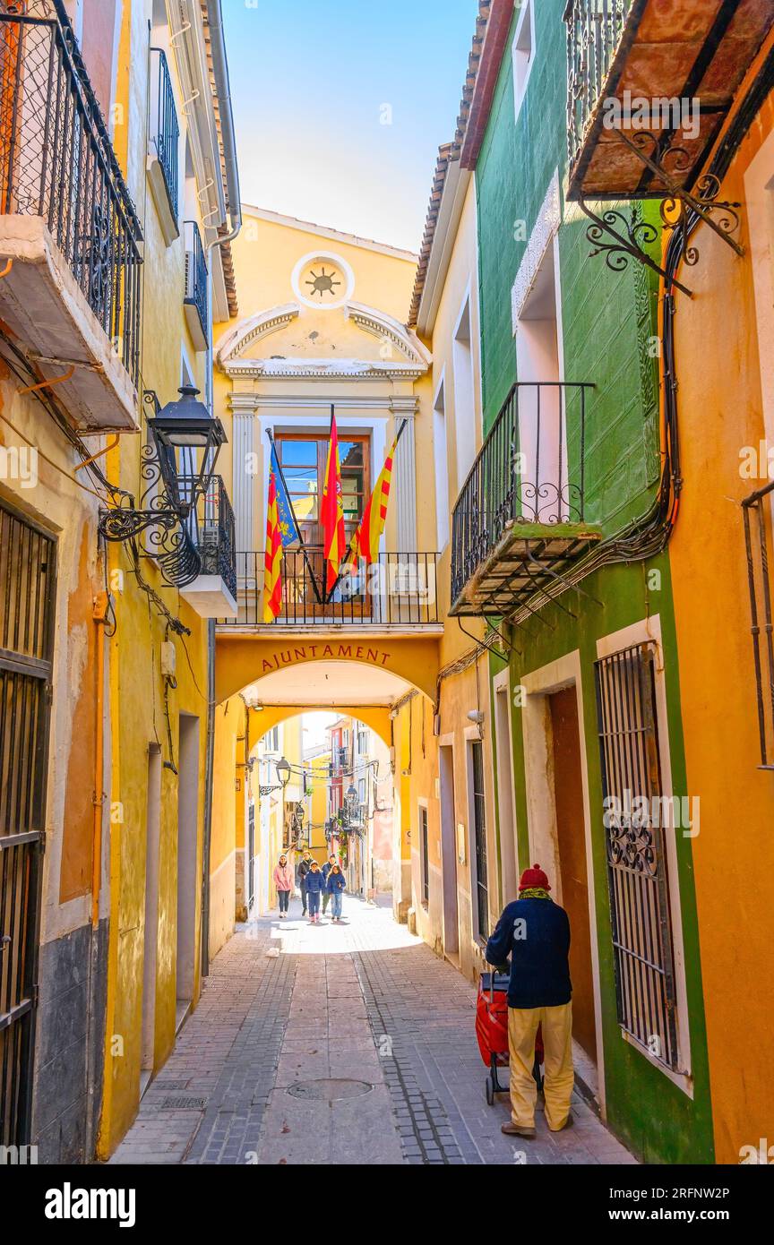 Villajoyosa, Spanien, Leute in einer engen Straße oder Gasse. Das Rathaus hat eine erhöhte Fußgängerbrücke. Andere Gebäude mit pastellfarbenen Wänden sind zu sehen Stockfoto