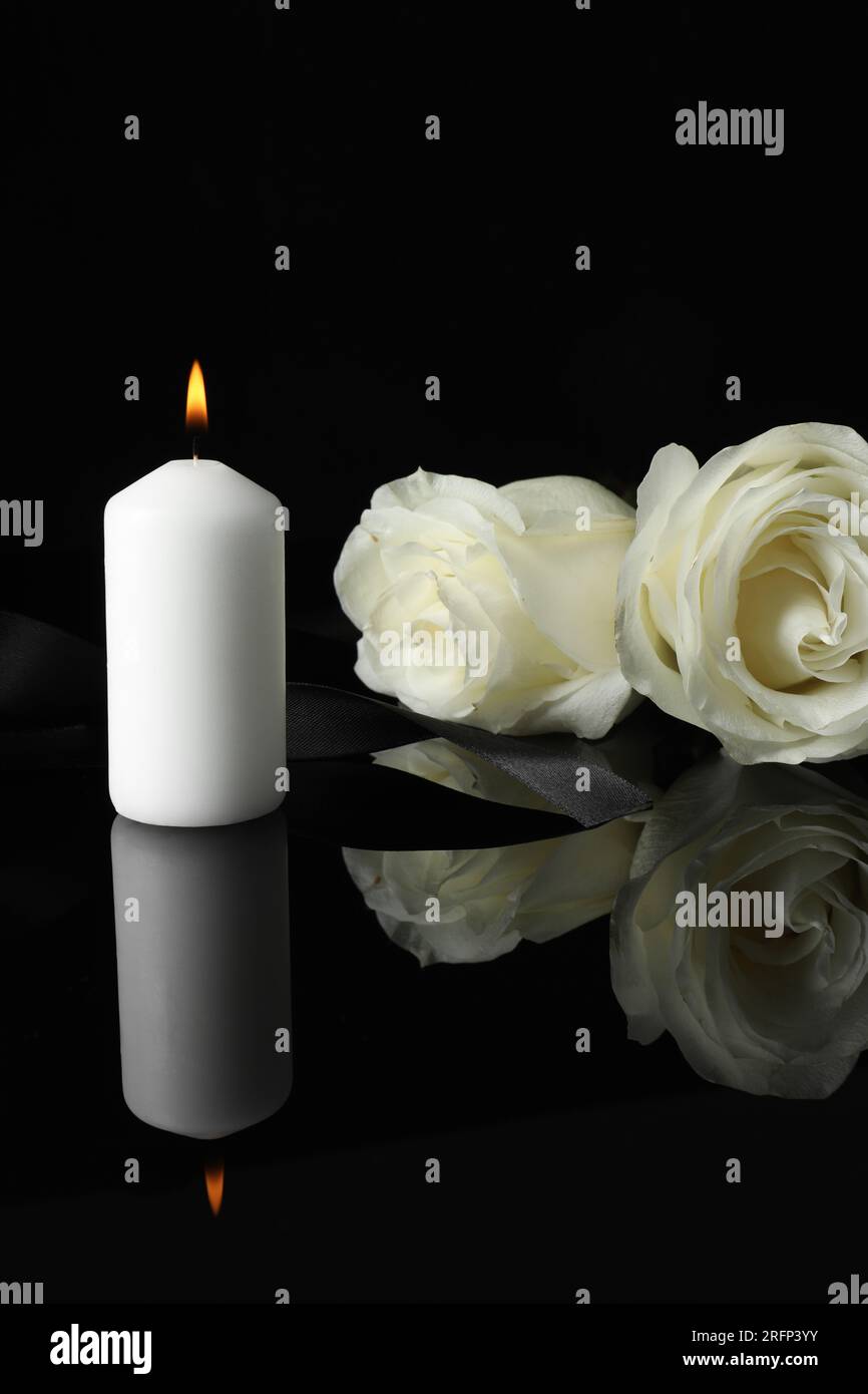 Brennende Kerze, weiße Rosen und Band auf schwarzer Spiegeloberfläche bei Dunkelheit, Platz für Text. Begräbnissymbole Stockfoto