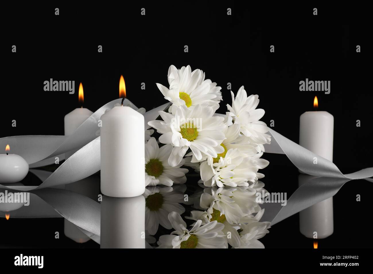 Weiße Chrysanthemblüten und brennende Kerzen auf schwarzer Spiegeloberfläche bei Dunkelheit. Begräbnissymbole Stockfoto