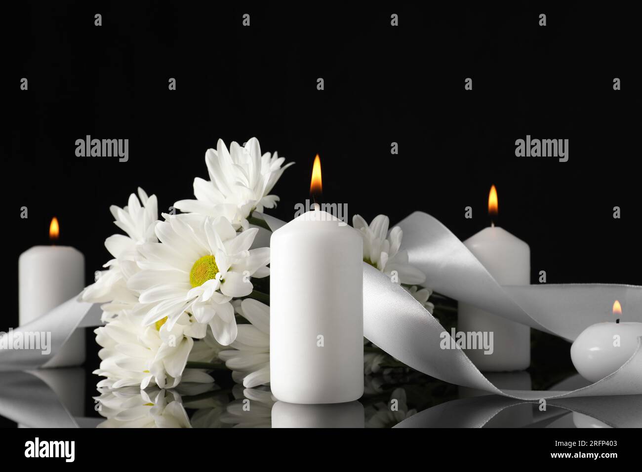 Weiße Chrysanthemblüten und brennende Kerzen auf schwarzer Spiegeloberfläche bei Dunkelheit. Begräbnissymbole Stockfoto