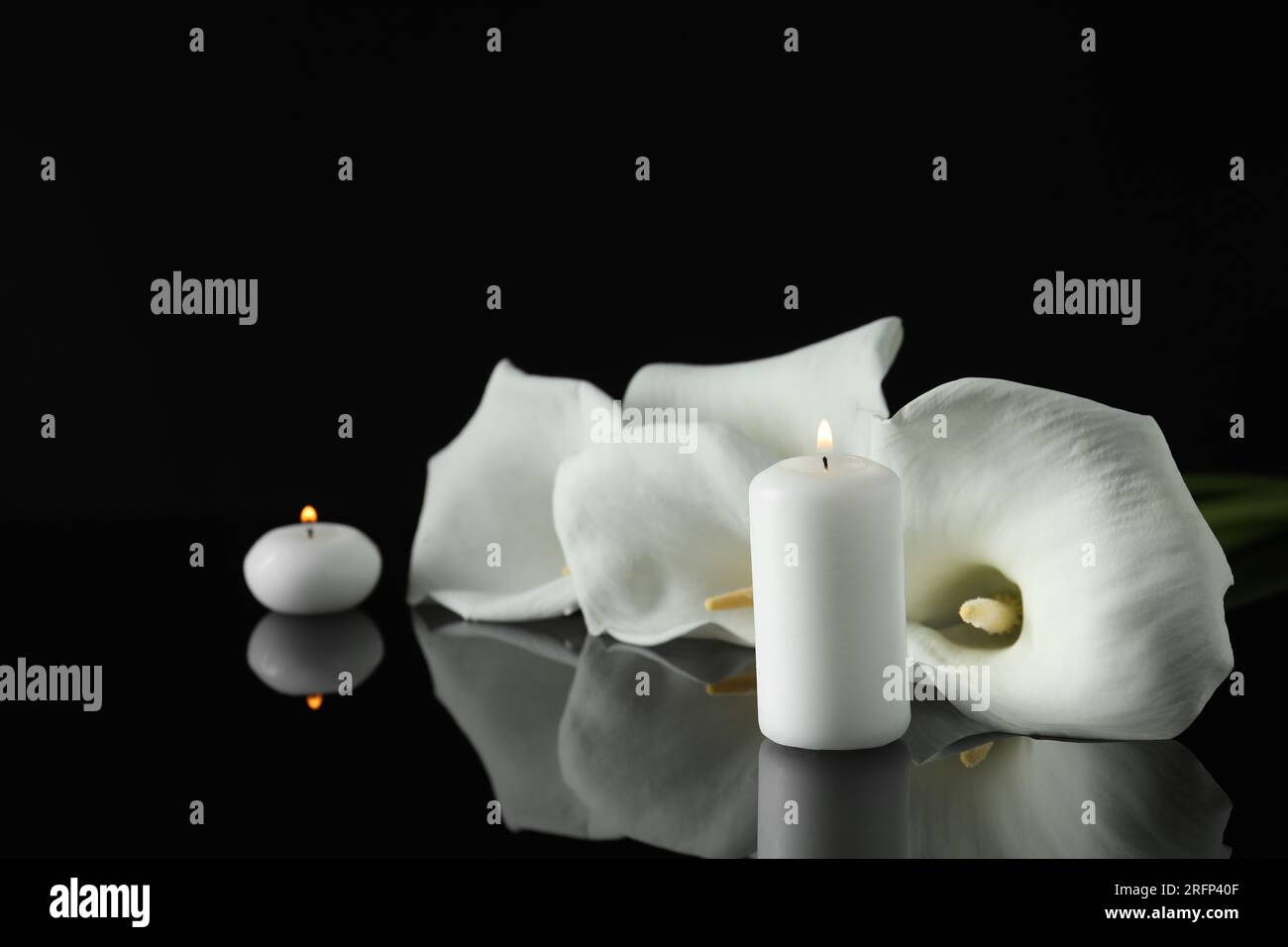 Weiße Calla Lilien Blumen und brennende Kerzen auf schwarzer Spiegeloberfläche in Dunkelheit, Platz für Text. Begräbnissymbole Stockfoto