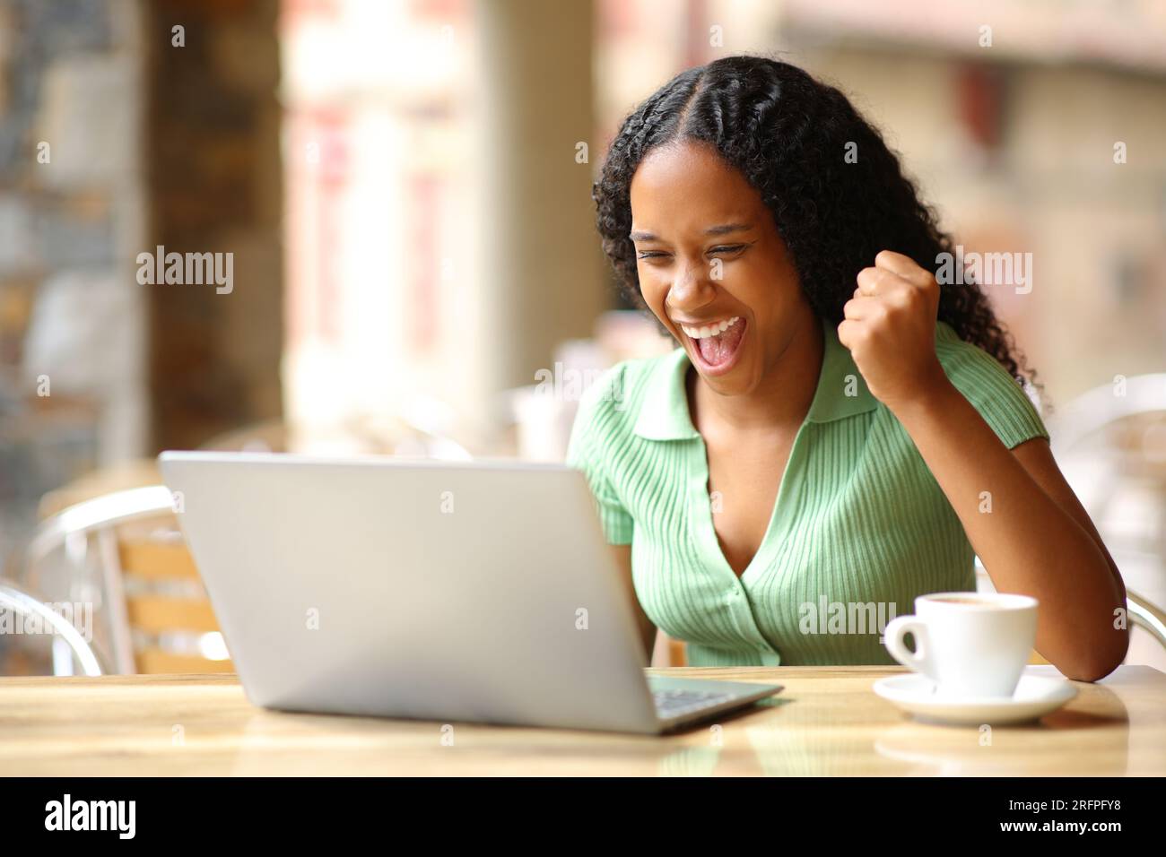 Aufgeregte schwarze Frau, die gute Nachrichten auf einem Laptop auf einer Barterrasse überprüft Stockfoto