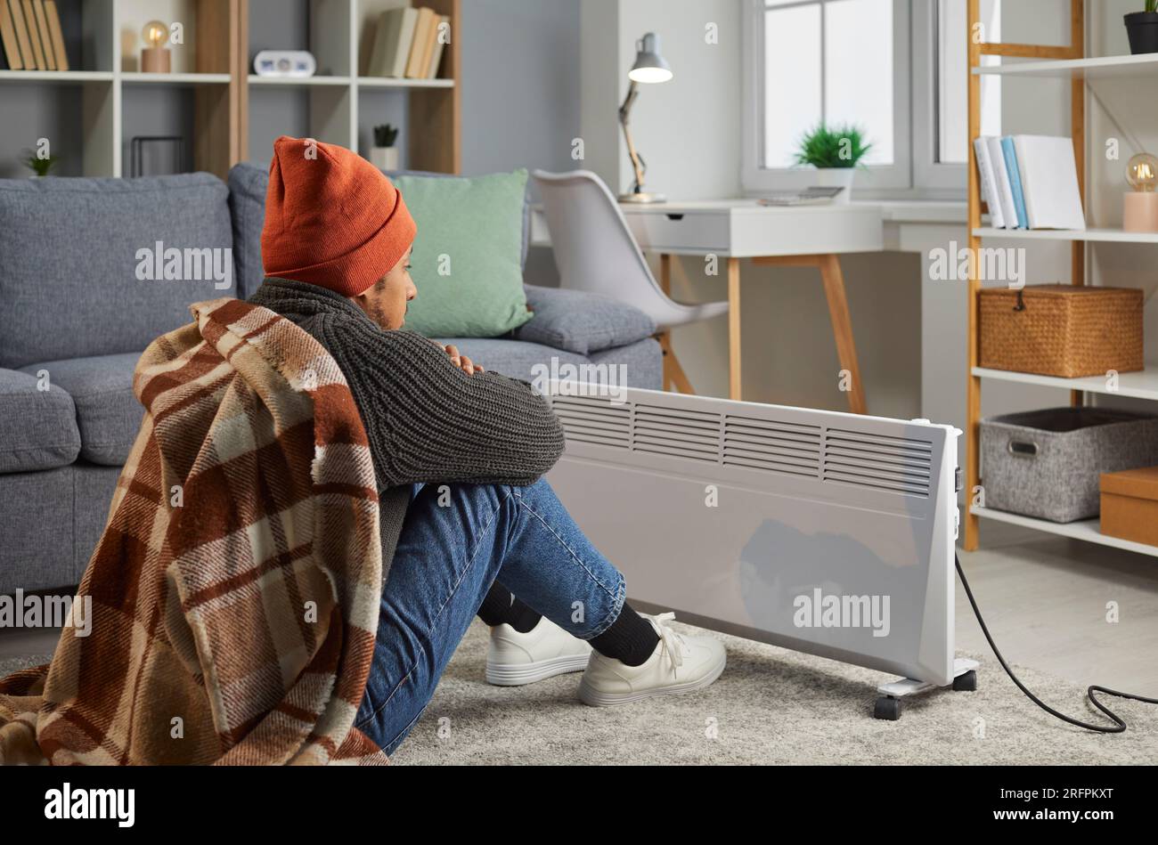 Junger gefrorener Mann, der sich zurücklehnt, in eine Decke eingewickelt, auf dem Boden zu Hause in der Nähe einer elektrischen Heizung. Stockfoto