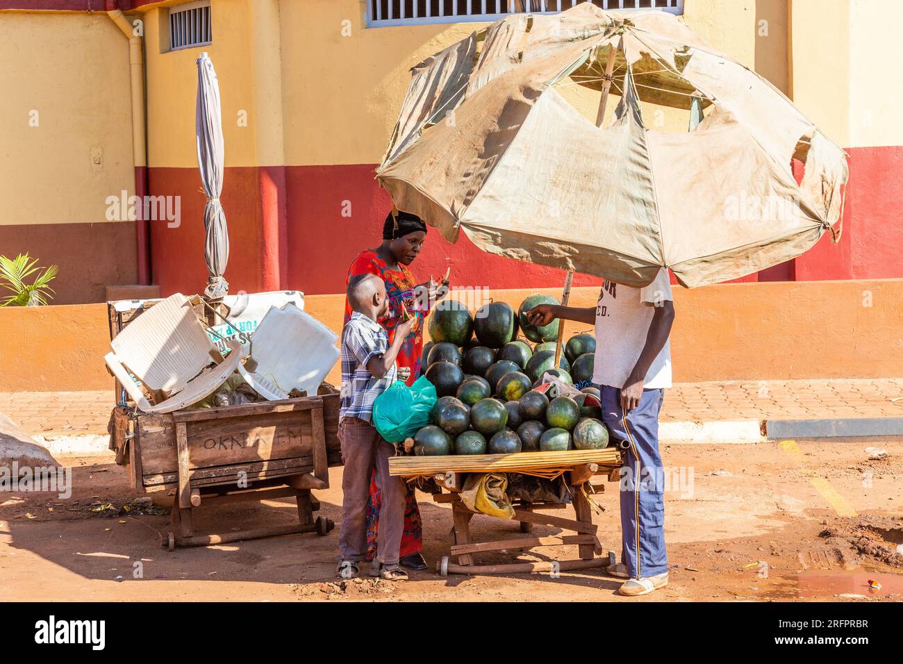 Geschützt unter einem Sonnenschirm verhandelt ein Wassermelonenverkäufer mit einer Frau und ihrem Sohn. Jinja, Uganda. Stockfoto