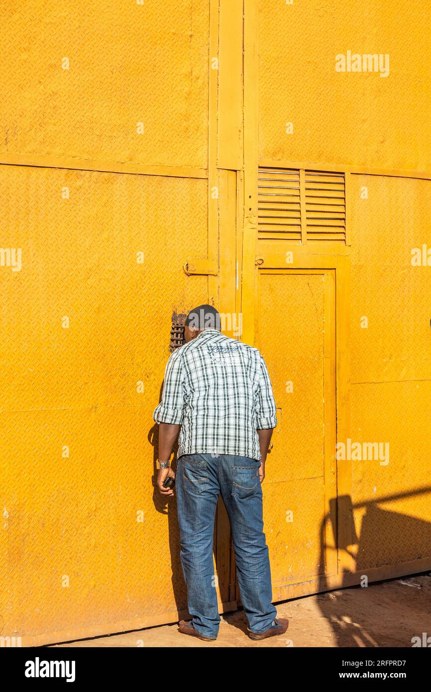 Ein Mann, der durch ein Wicket redete, durchbohrt in eine intensive gelbe Metalltür. Jinja, Uganda. Stockfoto