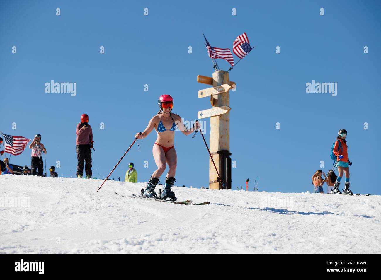 Mammoth Lakes, Kalifornien. 4. Juli 2023. An klaren Sommertagen kommt eine fitte Frau in einem Bikini und Helm vorbei an amerikanischen Flaggen im Mammoth Mountain Ski Resort. Stockfoto