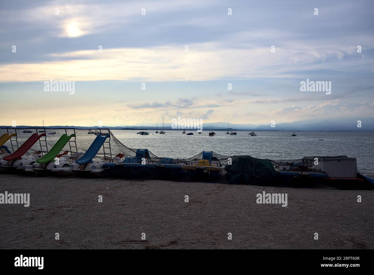 Strand auf einem See mit Paddelbooten und Menschen schwimmen auf einem bewölkten Sonnenuntergang mit einem Bergkamm in der Ferne Stockfoto