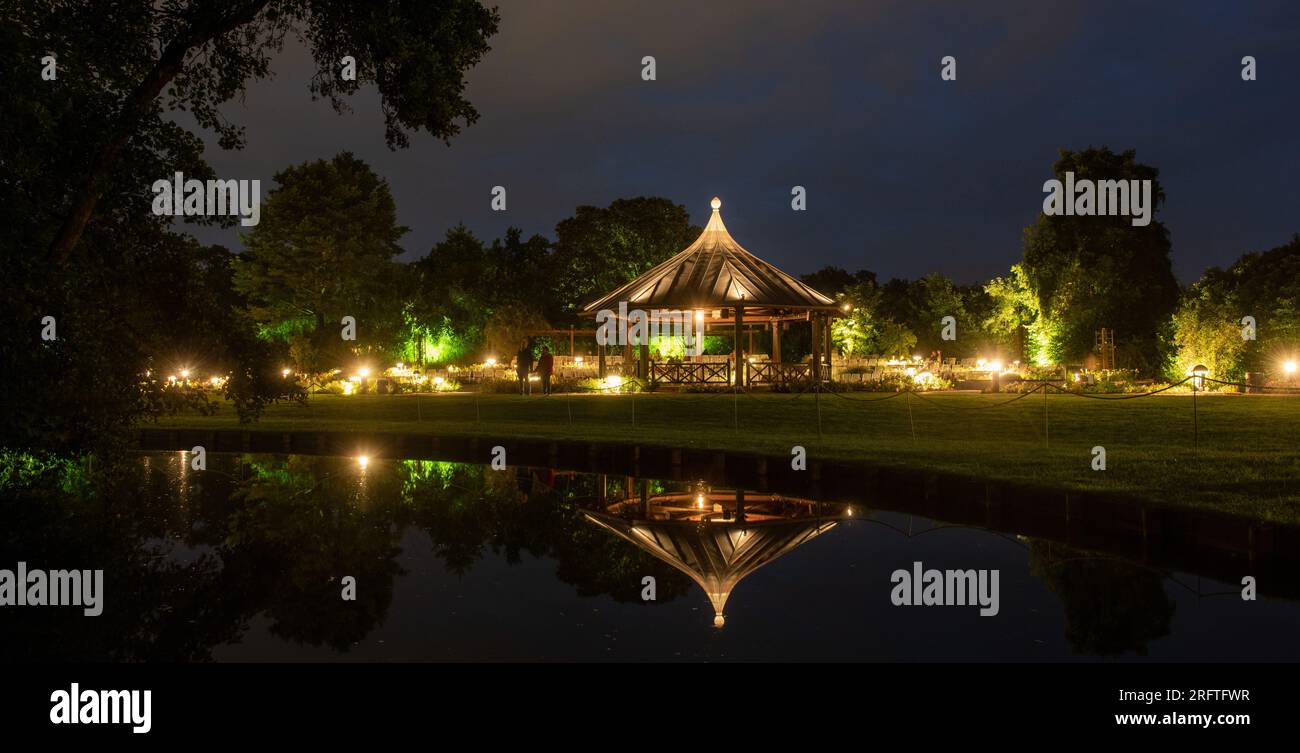 Augsburg, Deutschland. 05. Aug. 2023. Während der „Magie der Lichter“ stehen die Menschen im Botanischen Garten neben dem beleuchteten Rosenpavillon, der sich in einem Teich spiegelt. Jeden Samstag, bis zum 13. August, gehen im Botanischen Garten unzählige Lichter in der Dämmerung an. Zu diesem Zweck ist der Botanische Garten bis Mitternacht geöffnet. Kredit: Stefan Puchner/dpa/Alamy Live News Stockfoto