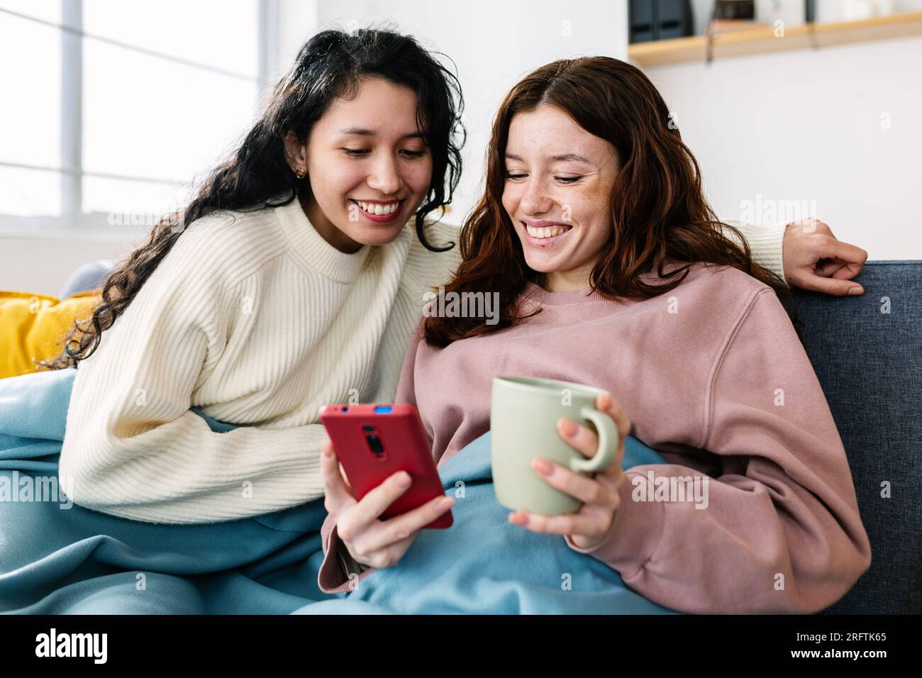 Glückliche junge Freundinnen, die ihr Handy benutzen und zu Hause auf dem Sofa sitzen Stockfoto