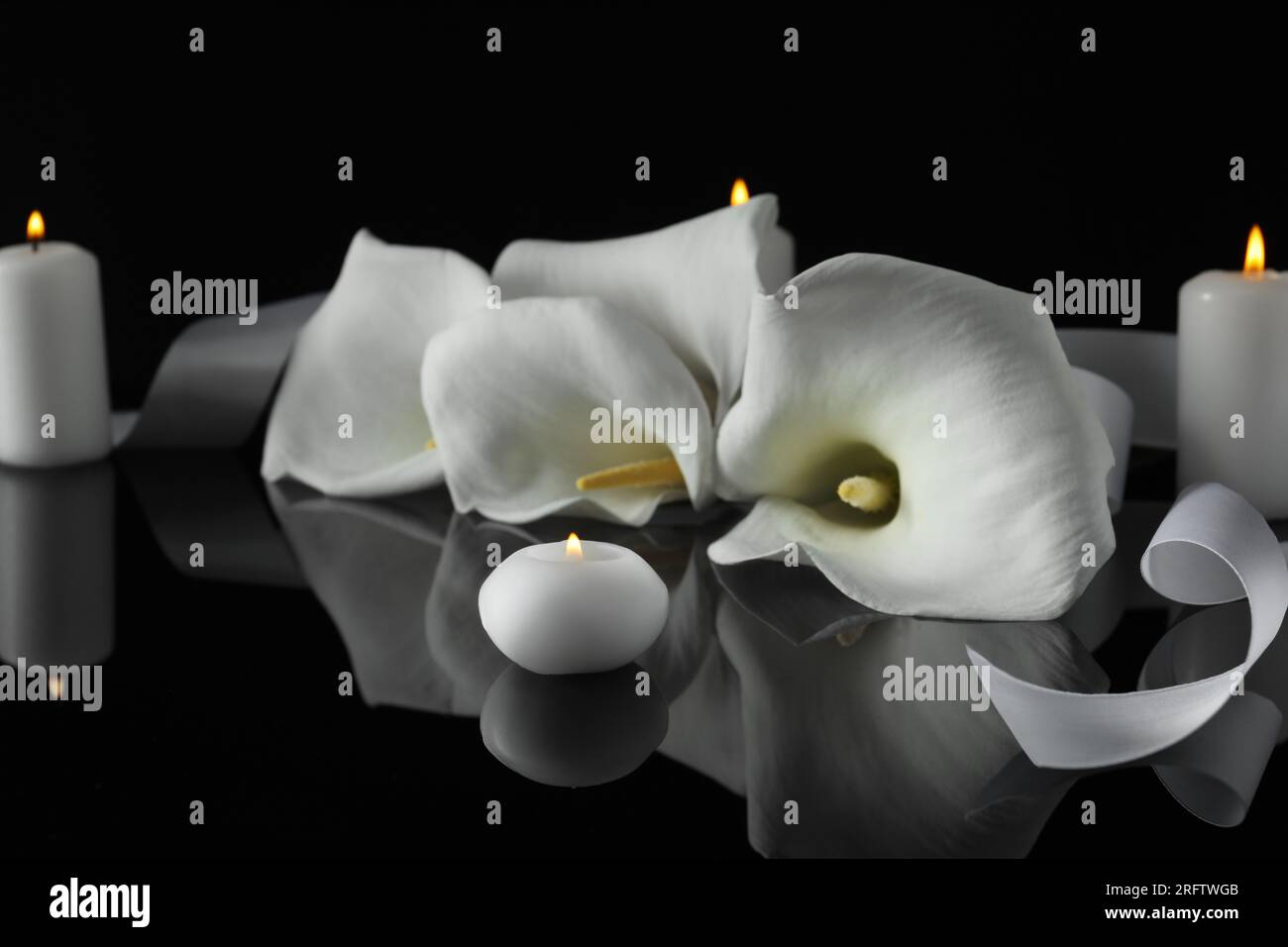 Brennende Kerzen und weiße Calla-Lilien-Blumen auf schwarzer Spiegeloberfläche in Dunkelheit, Nahaufnahme. Begräbnissymbole Stockfoto