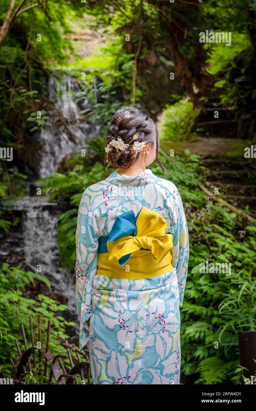 Rückansicht einer Frau mit japanischem Yukata-Sommerkimono, die auf der Straße neben dem Waldwasserfall in der Natur spaziert. Kyoto, Japan. Stockfoto