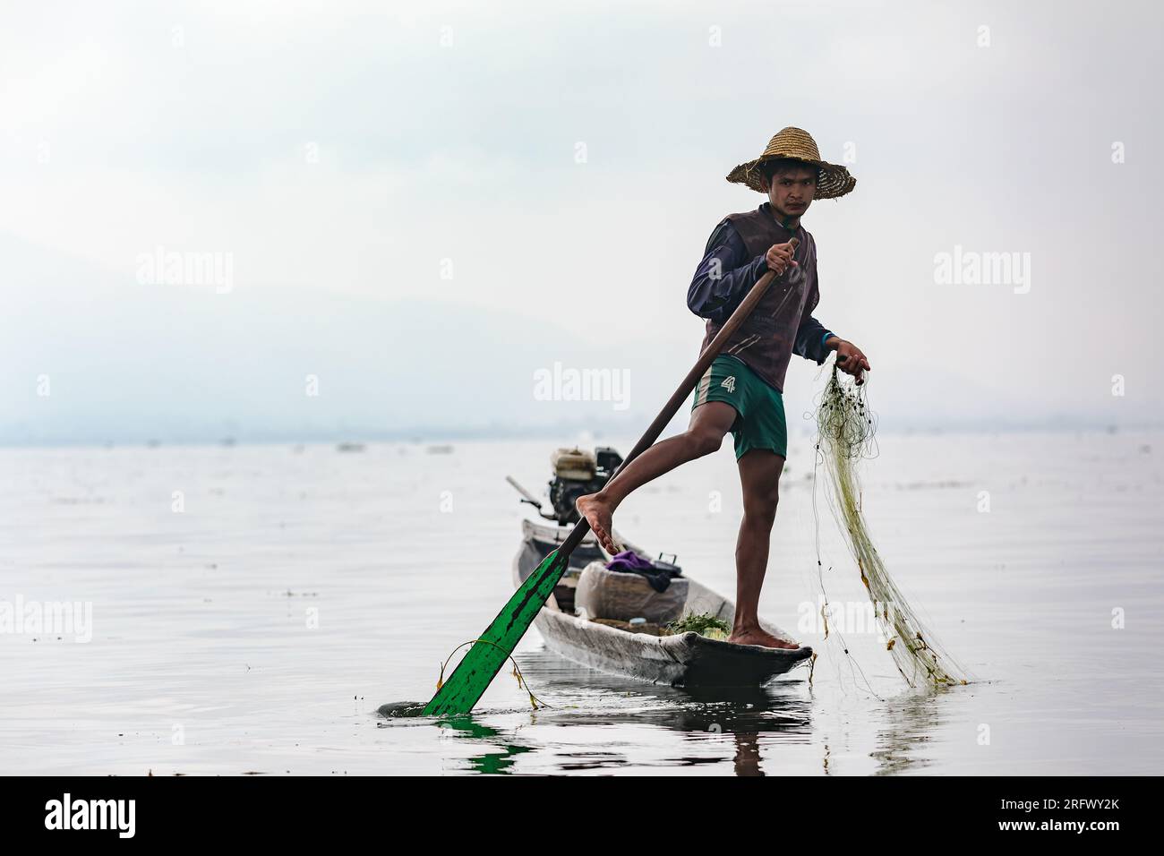 Ein Fischer steht auf einem Bein am Inle Lake und wickelt ein Bein um das Ruder des langen Boots, sodass seine Hände frei sind, um Fische zu fangen, Myanmar Stockfoto