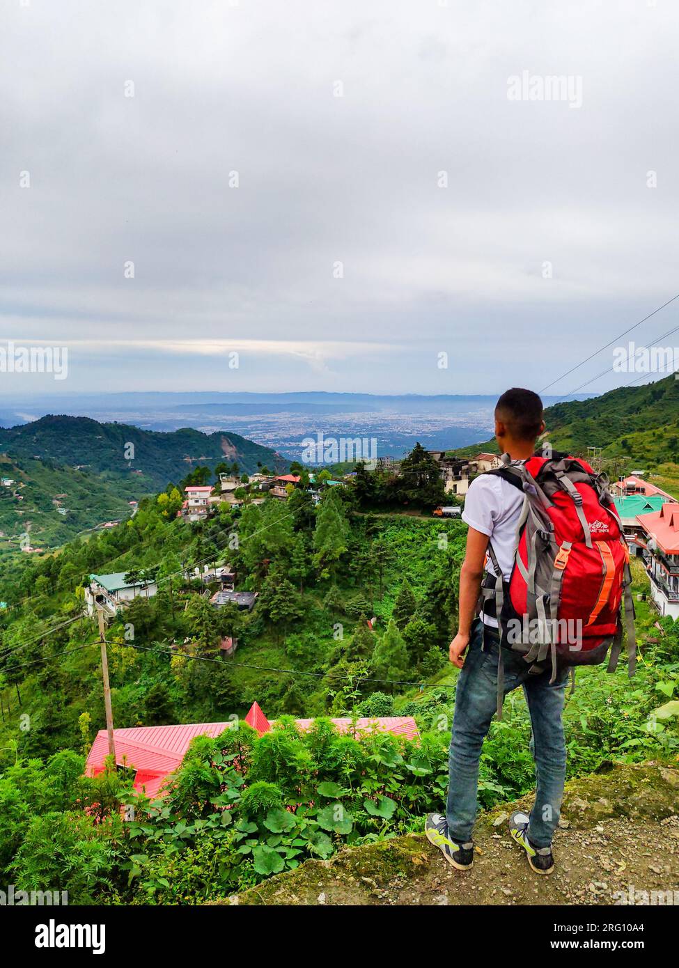 Oktober 14. 2022, Uttarakhand Indien. Junger Mann mit einem großen roten Rucksack, der das Doon Valley von Mussoorie Hills, Uttarakhand, Indien, beobachtet. Ruhiger Hügel Stockfoto