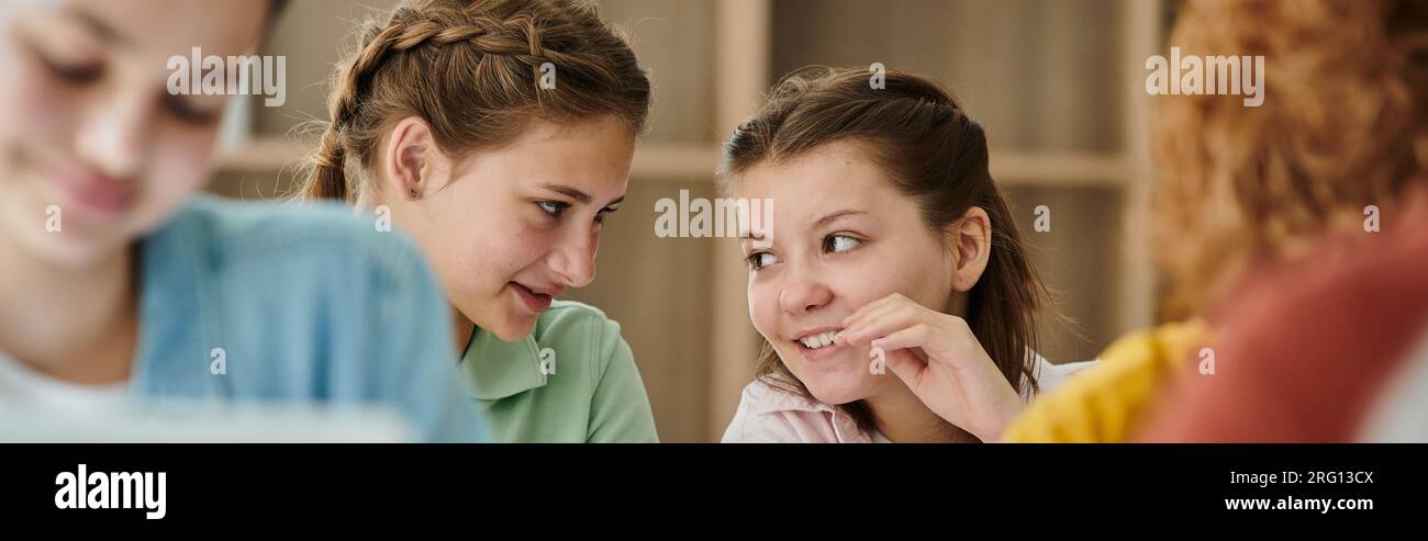 Lächelndes Schulmädchen, das mit einem Teenager-Freund während des Unterrichts in der Unschärfe-Klasse in der Schule spricht, Banner Stockfoto