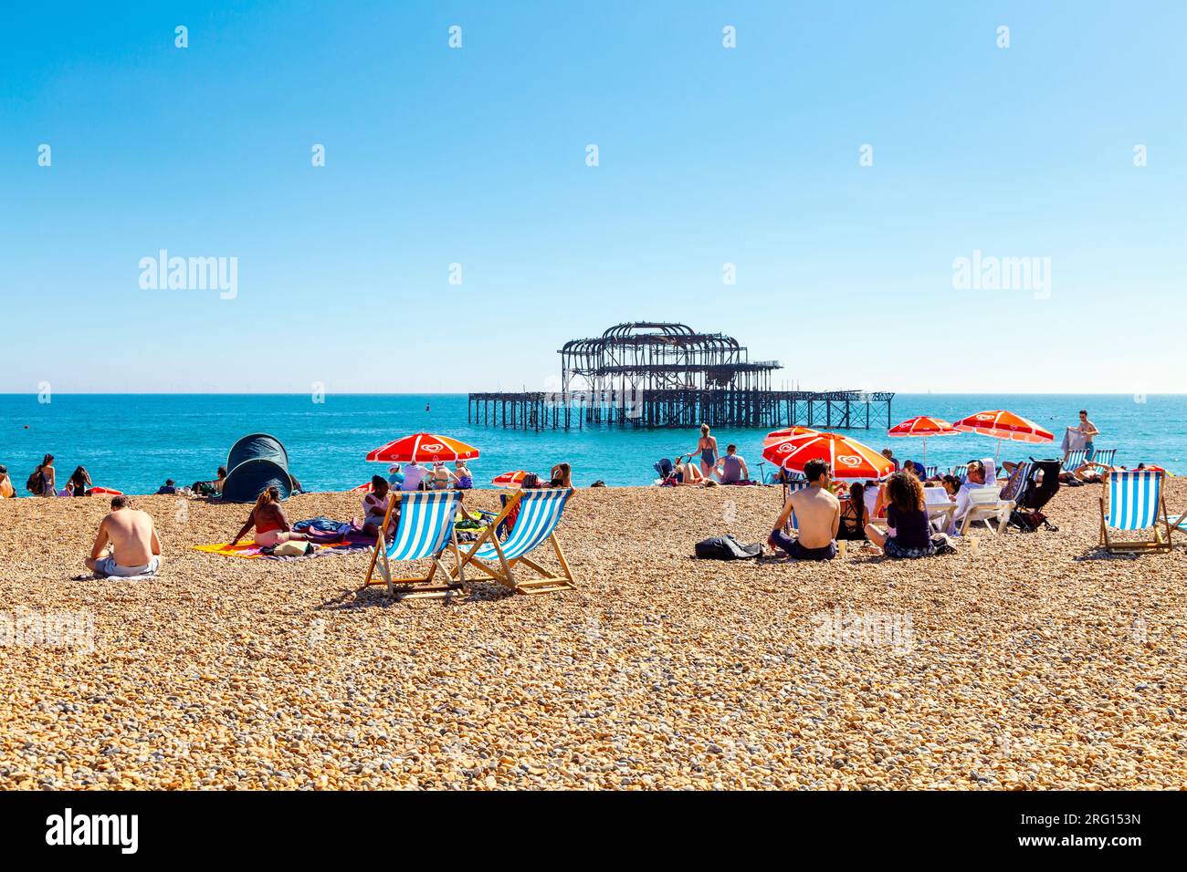 Leute, die an einem heißen Sommertag am Strand sitzen, mit dem abgebrannten West Pier im Hintergrund, Brighton, Großbritannien Stockfoto
