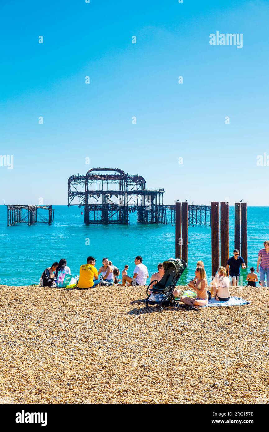Leute, die an einem heißen Sommertag am Strand sitzen, mit dem abgebrannten West Pier im Hintergrund, Brighton, Großbritannien Stockfoto