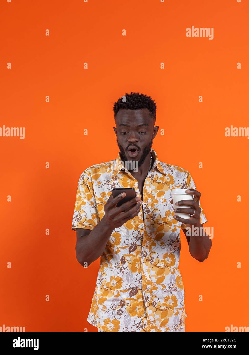 Afroamerikanischer Mann hält Smartphone und nimmt Selbstporträt mit offenem Mund auf, während er Kartonglas auf orangefarbenem Hintergrund hält Stockfoto