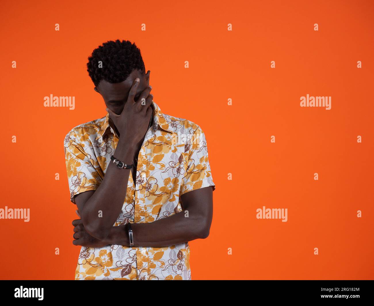 Deprimierter junger afroamerikanischer Mann in orangefarbenem bedrucktem Kurzarmhemd und geschlossenen Augen, während er mit gekreuzten Armen stand und sich das Gesicht in der Hand versteckte Stockfoto