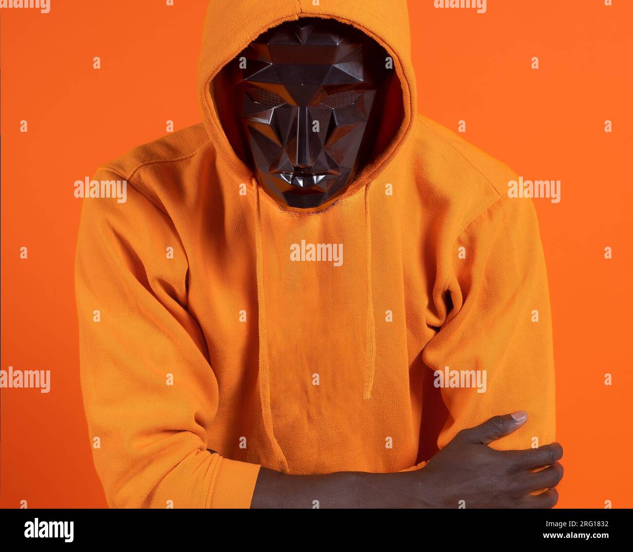 Anonyme schwarze Person in schwarz verschwommener Gesichtsmaske und orangefarbener Kapuzenpullover mit gekreuzter Hand vor orangefarbenem Studiohintergrund Stockfoto