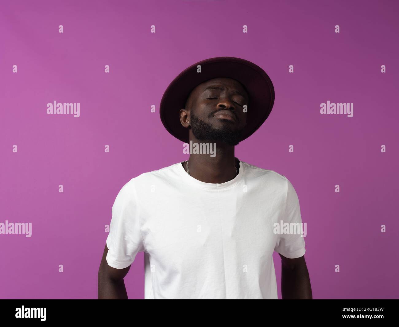 Erschöpfter junger afroamerikanischer Mann mit Hut und weißem Hemd mit geschlossenen Augen, während er allein in deprimierter und trauernder Stimmung vor violettem Hintergrund steht Stockfoto
