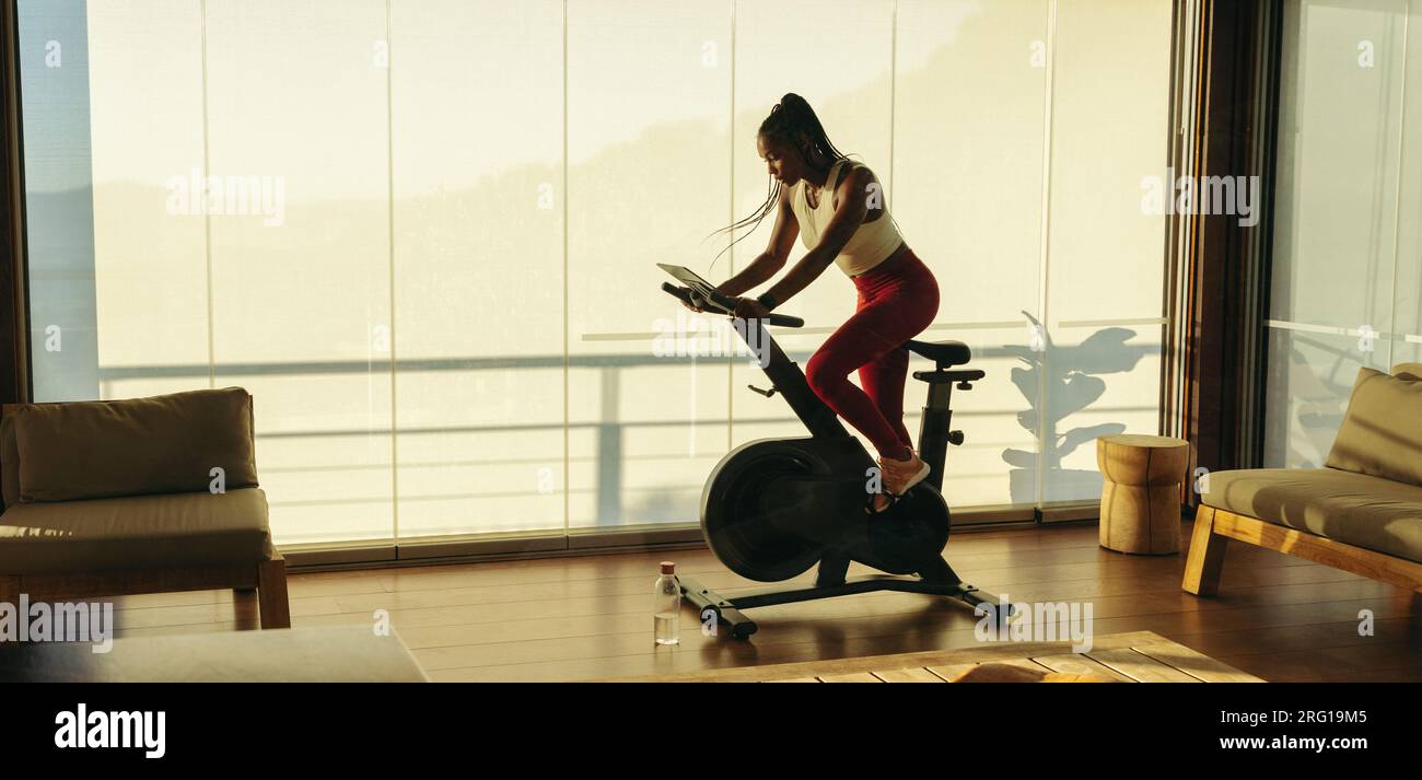 Sportliche schwarze Frau, die sich zu Hause zu ihrem Fitnesstraining verpflichtet und mit einem intelligenten Fitness-Bike Cardio-Training durchführt. Junge Frau, die hart arbeitet, um ihren Zustand zu erhalten Stockfoto