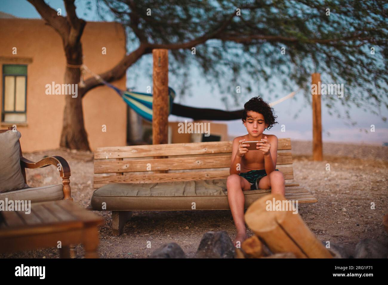 Ein kleiner Junge, der mit seinem Smartphone spielt und auf einer Bank in der Wüste sitzt Stockfoto