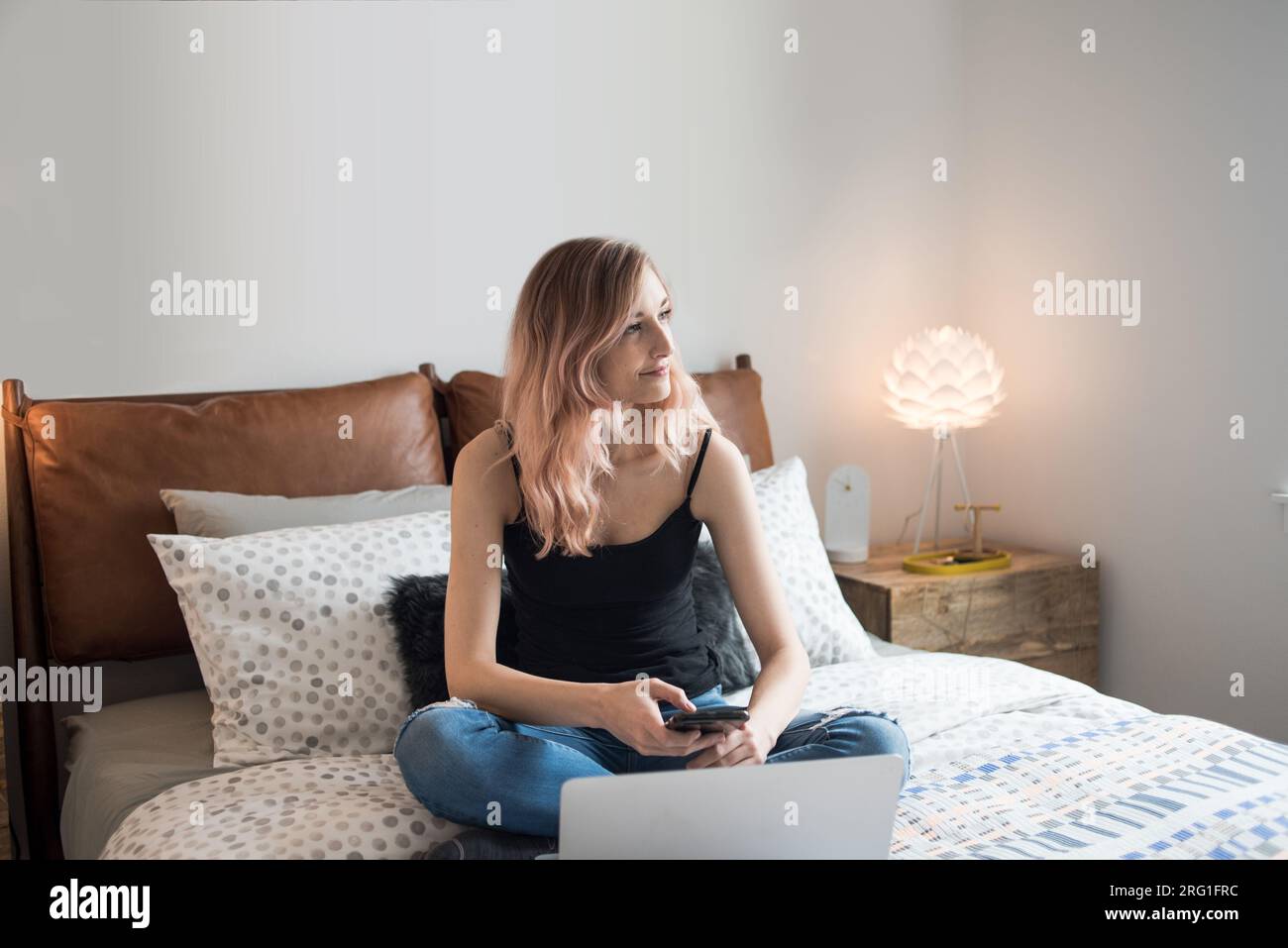 Eine Frau, die auf dem Bett sitzt und von der Kamera schaut und ein Smartphone hält Stockfoto