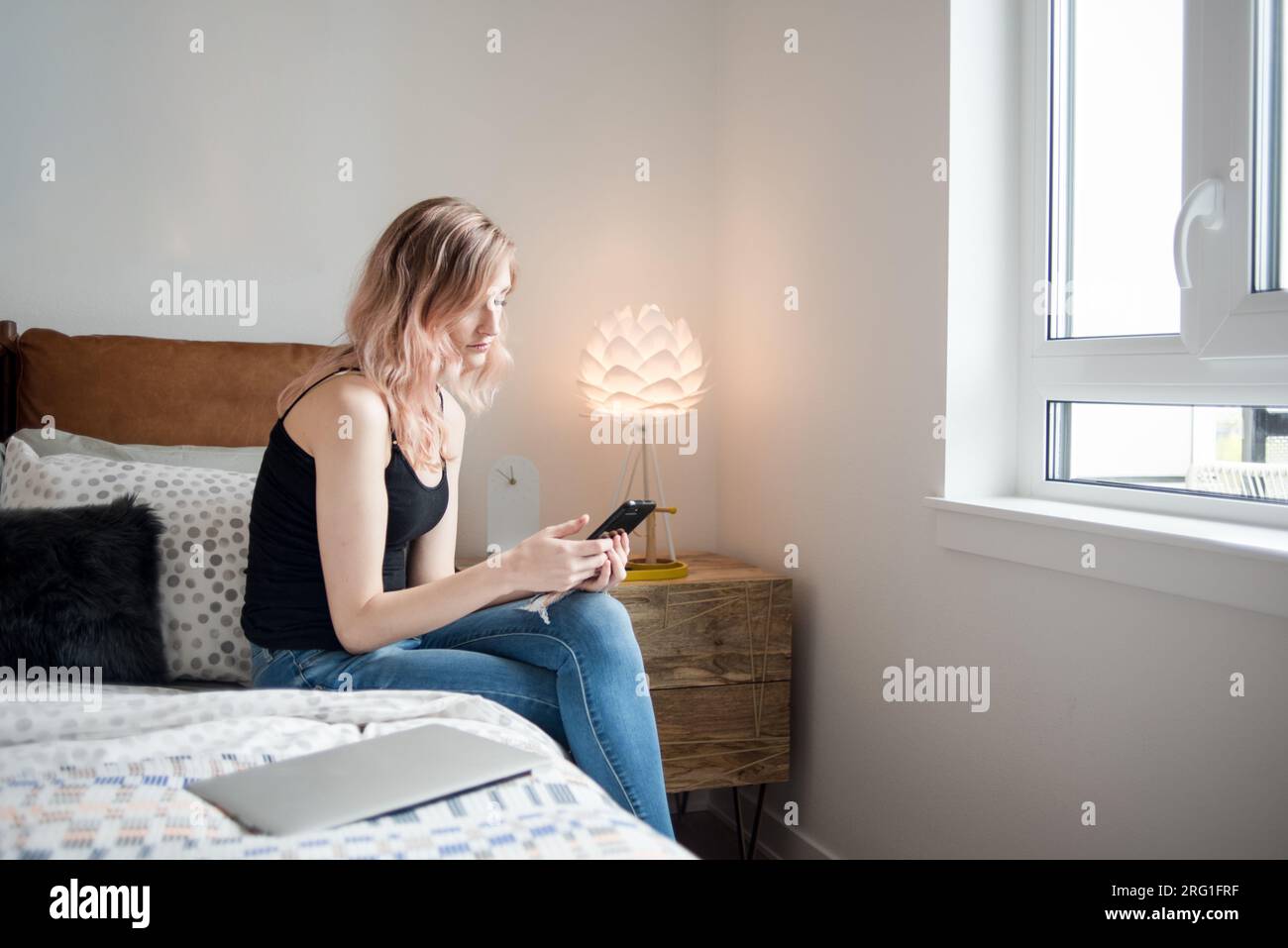 Frau, die mit Smartphone und Laptop neben dem Bett sitzt Stockfoto