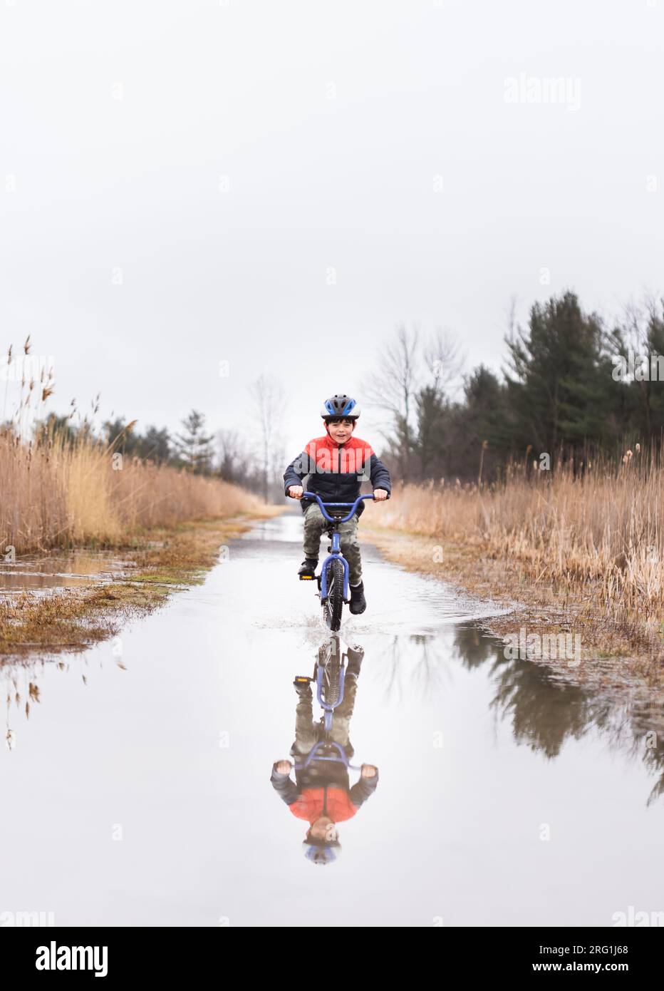 Ein kleiner Junge, der mit dem Fahrrad durch eine Pfütze auf einem überfluteten Pfad fährt. Stockfoto