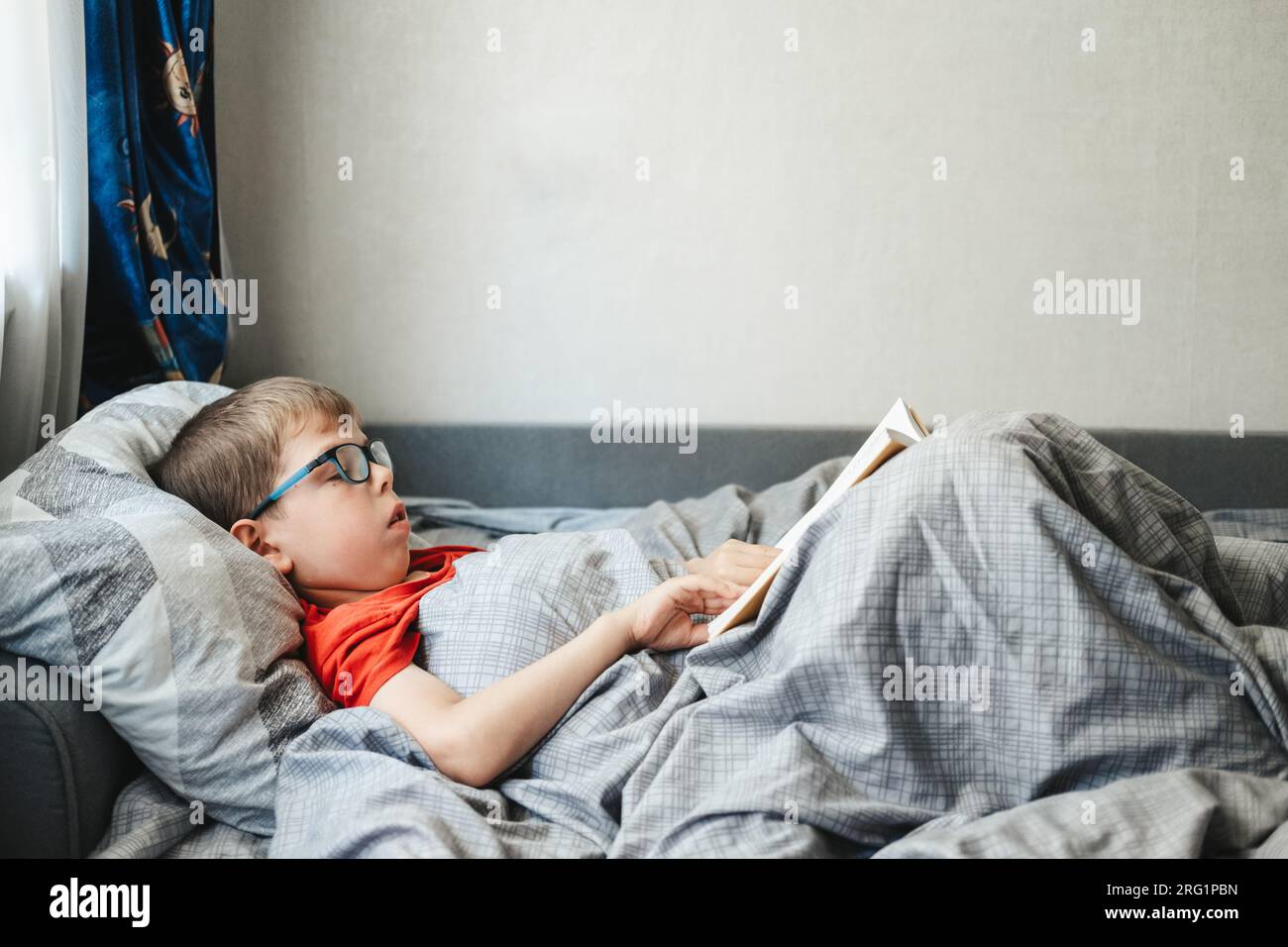 Ein Junge mit Brille liegt auf einem Bett unter einer Decke und liest ein Buch. Ein Kind im Grundschulalter liest ein Buch in seinem Bett. Stockfoto