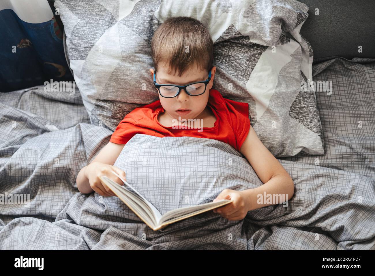 Ein Junge mit Brille liegt auf einem Bett unter einer Decke und liest ein Buch, Blick von oben. Ein Kind im Grundschulalter liest ein Buch in seinem Bett. Stockfoto