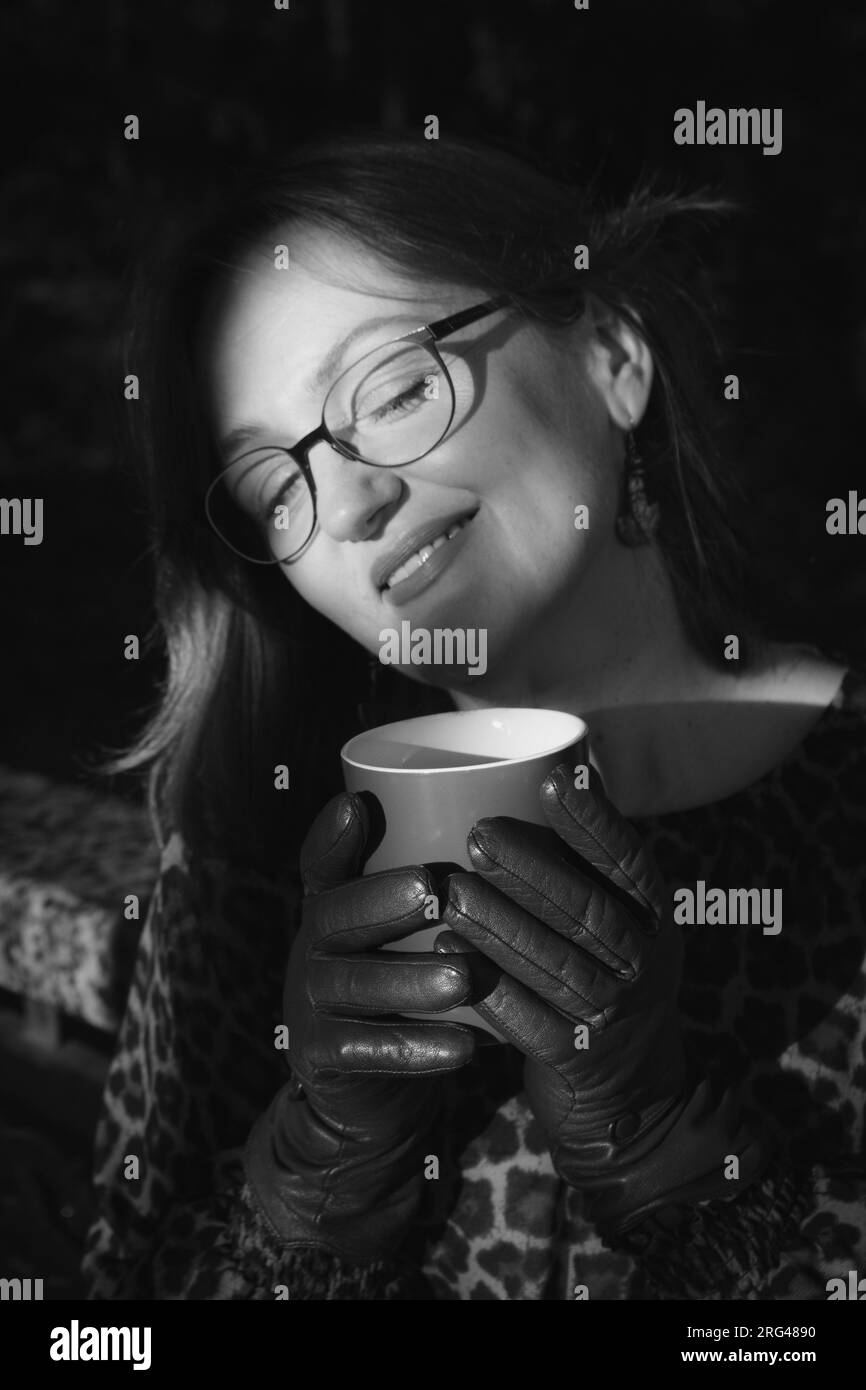 Schwarz-Weiß-Porträt einer lächelnden Frau mit geschlossenen Augen. Reife Frau im Morgensonnenlicht mit Kaffeetasse, schwarz-weiß. Konzept des Selbstwertgefühls. Stockfoto
