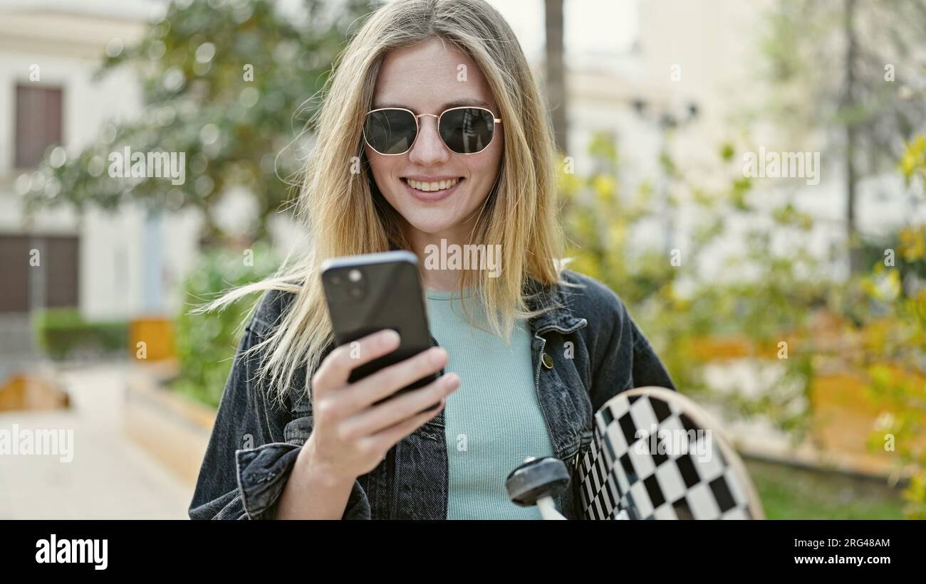 Junge blonde Frau, die im Park mit dem Smartphone Schlittschuh hält Stockfoto