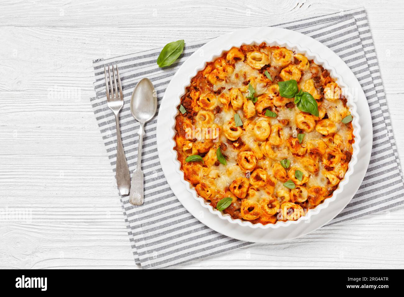 Tortellini gebacken mit Mozzarella-Käse, Tomatensoße und Rinderhack in weißer Backform auf weißem Holztisch mit Löffel und Gabel, horizontale Ansicht Stockfoto