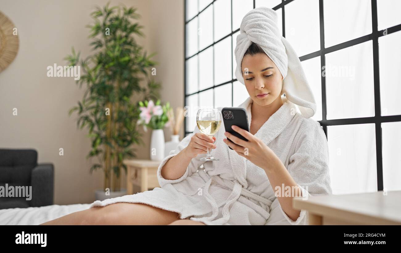 Junge, wunderschöne hispanische Frau im Bademantel, die Champagner mit dem Smartphone im Schlafzimmer trinkt Stockfoto