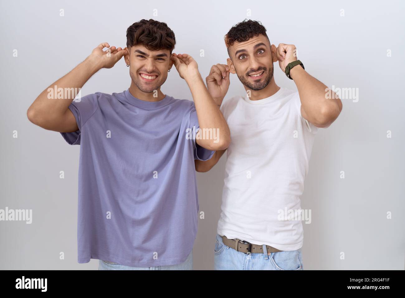 Homosexuelles schwules Paar, das über weißem Hintergrund steht und lächelt, mit Fingern an den Ohren zieht, lustige Geste. Audition-Problem Stockfoto