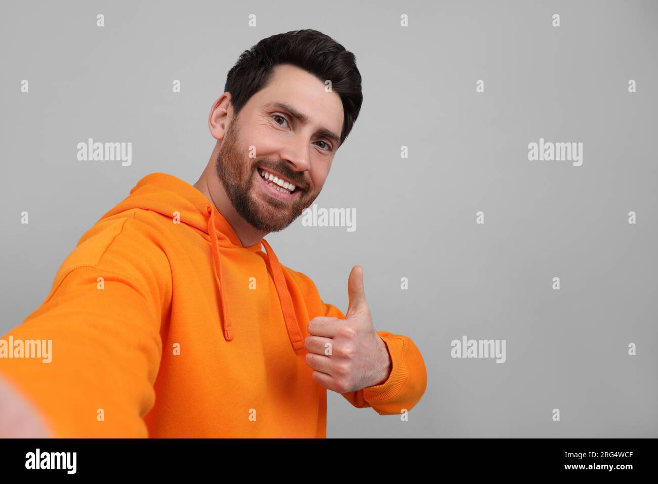 Ein lächelnder Mann macht Selfie und zeigt Daumen nach oben auf grauem Hintergrund, Platz für Text Stockfoto