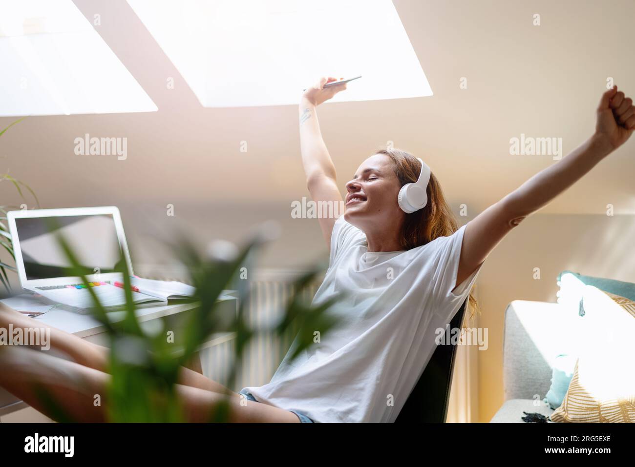 Glückliche junge Frau mit Kopfhörern hört gern ihre persönliche Playlist, während sie zu Hause am Schreibtisch sitzt und sich ausstreckt Stockfoto