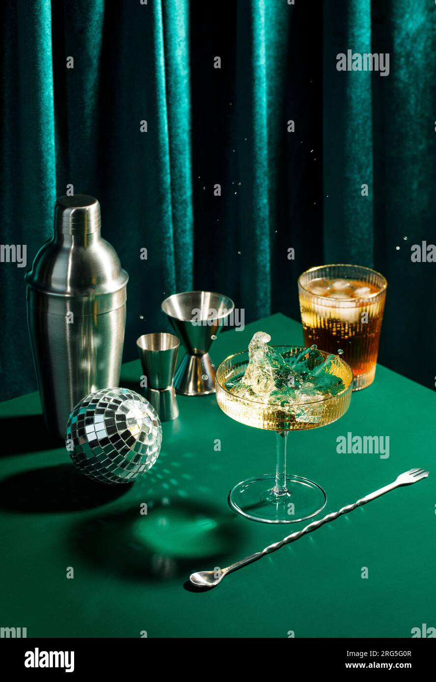 Planschen Sie in Champagnerglas, Cocktailglas, Shaker, Cocktaillöffel und Jigger auf einem dunkelgrünen Tisch in Disco-Club-Atmosphäre Stockfoto