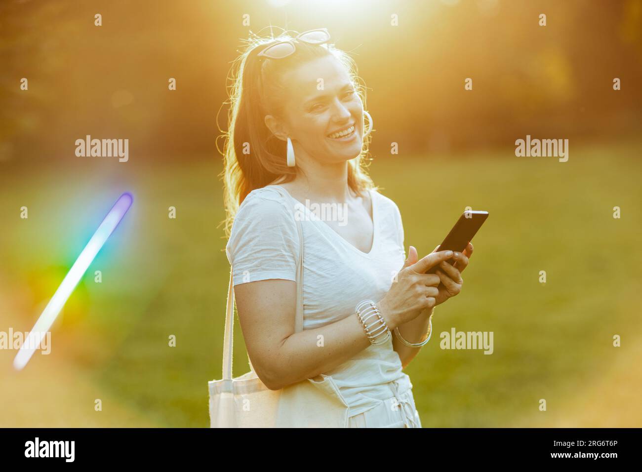 Sommerzeit. Eine glückliche, stilvolle Frau in einem weißen Hemd, die auf der Wiese im Stadtpark ein Smartphone benutzt. Stockfoto
