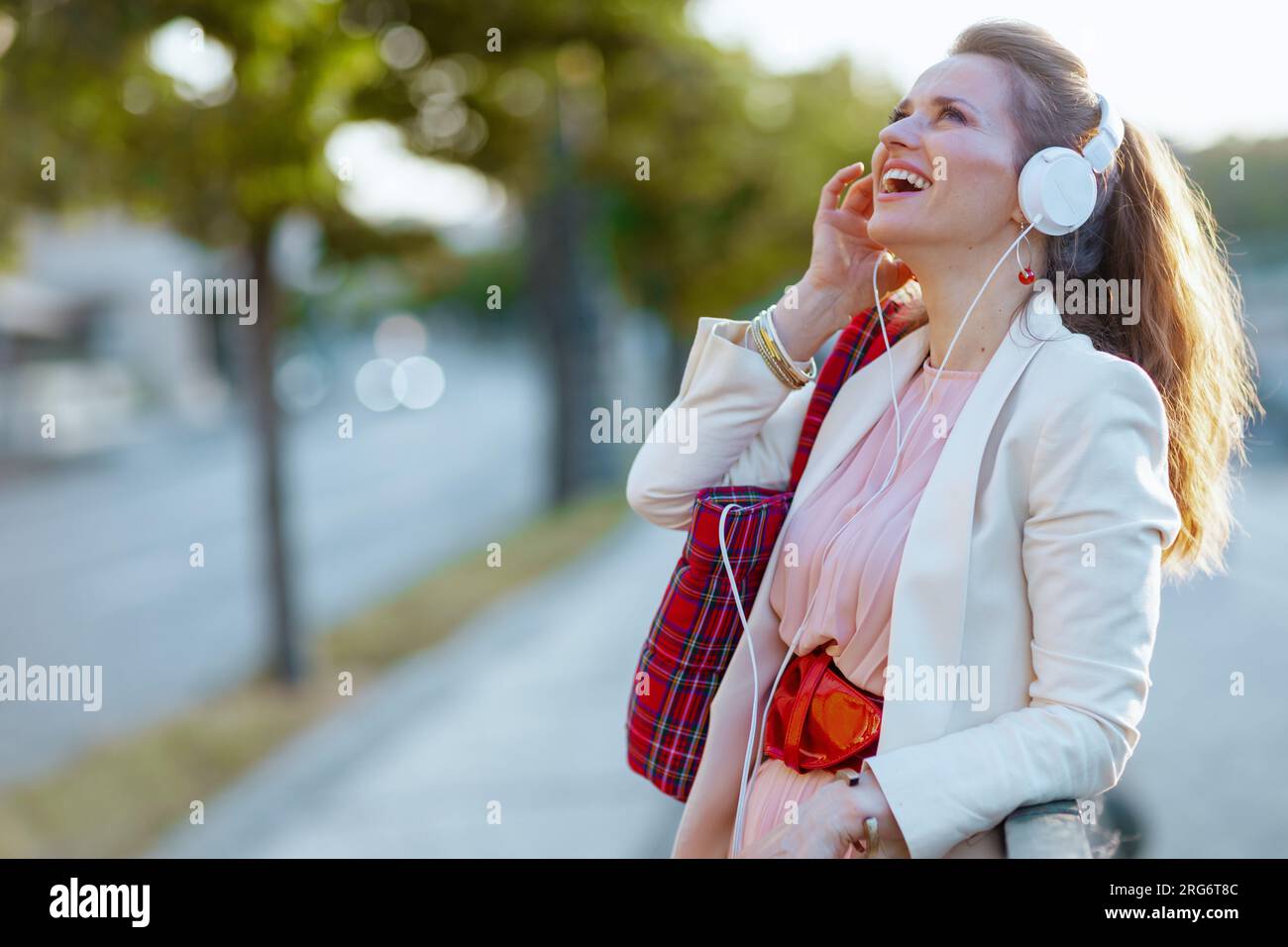 Fröhliche, stilvolle Frau in pinkfarbenem Kleid und weißer Jacke in der Stadt mit roter Tasche, die Musik über Kopfhörer hört. Stockfoto