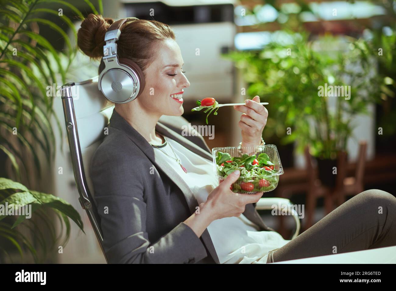 Nachhaltiger Arbeitsplatz. Lächelnde, moderne Eigentümerin eines kleinen Unternehmens mittleren Alters in einem grauen Business-Anzug in einem modernen grünen Büro, die Salat isst und zuhört Stockfoto