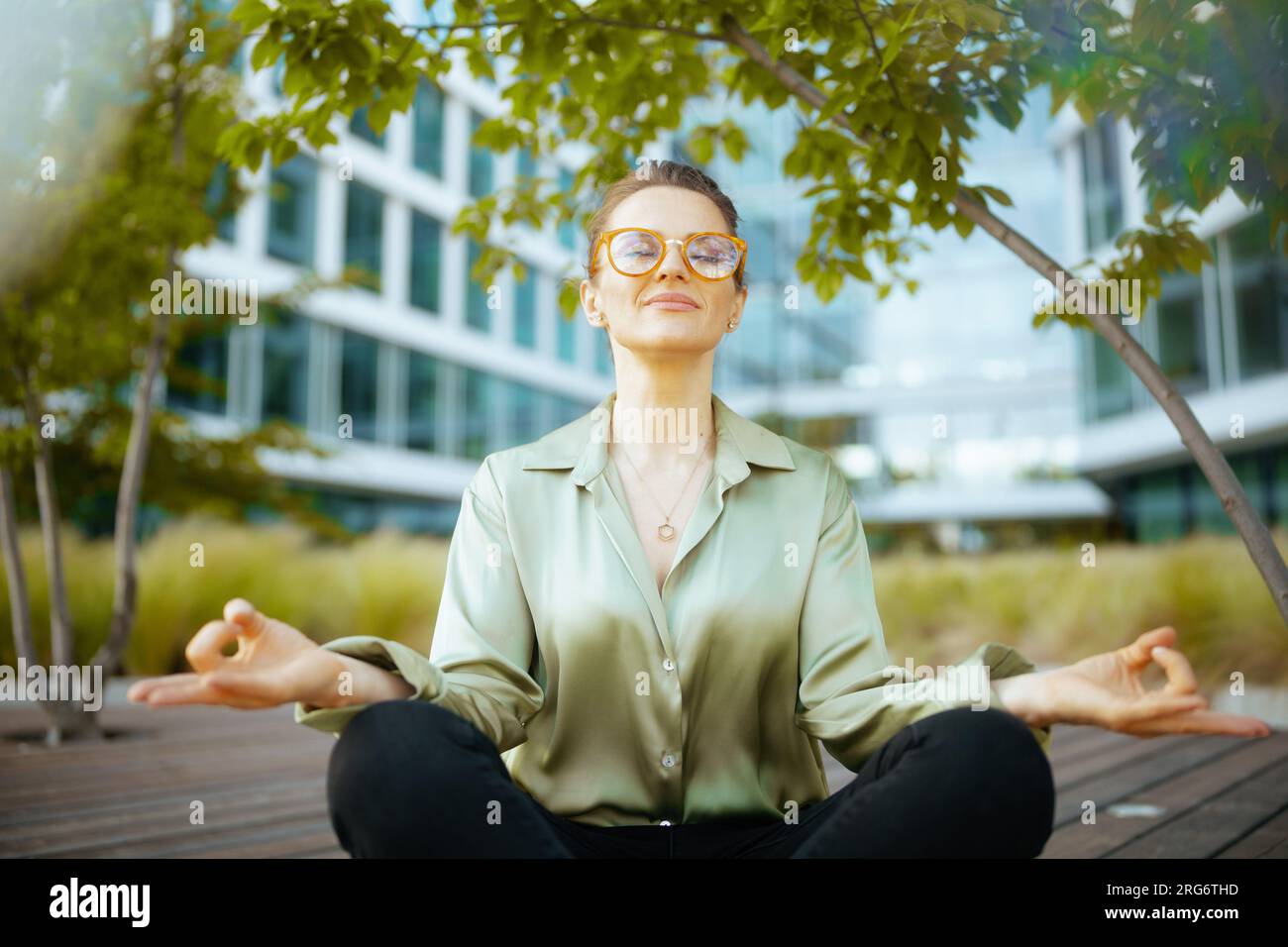 Entspannte, moderne Mitarbeiterin mittleren Alters in der Nähe des Business Centers mit grüner Bluse und Brille beim Meditieren. Stockfoto