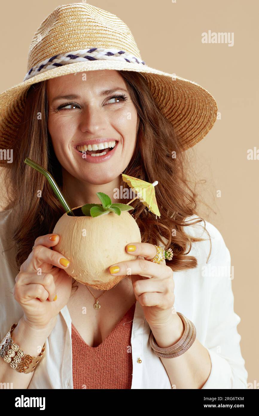 Strandurlaub. Fröhliche, trendige Frau in weißer Bluse und Shorts isoliert auf Beige mit Kokosnuss-Cocktail und Sommerhut. Stockfoto Strandurlaub. Fröhliche, trendige Frau in weißer Bluse und Shorts isoliert auf Beige mit Kokosnuss-Cocktail und Sommerhut. Stockfoto