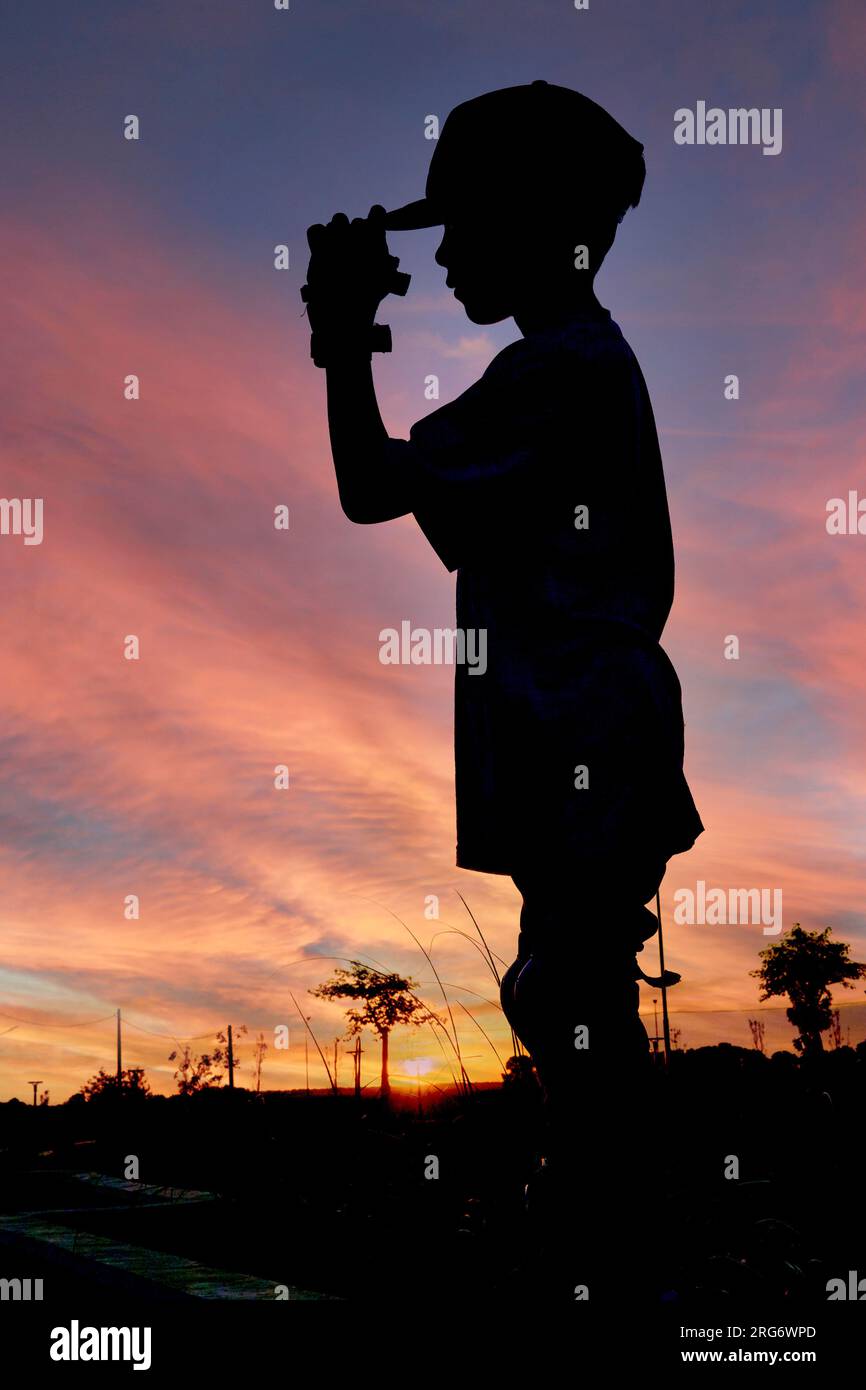 Hintergrundbeleuchtetes Foto eines Jungen, der im Profil steht und sein Visier hält, mit Sonnenuntergang im Hintergrund Stockfoto