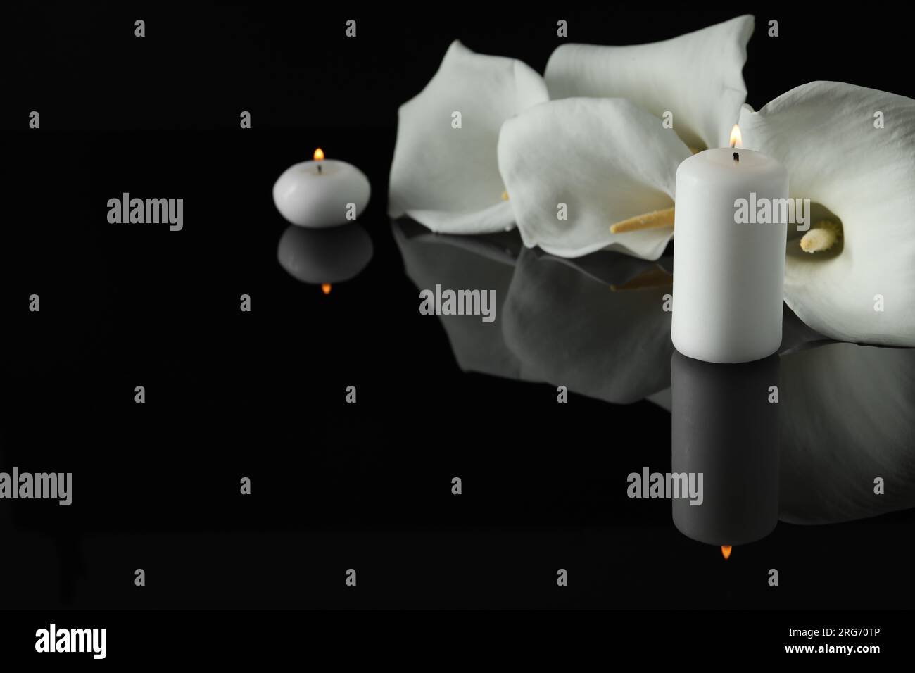 Weiße Calla Lilien Blumen und brennende Kerzen auf schwarzer Spiegeloberfläche in Dunkelheit, Platz für Text. Begräbnissymbole Stockfoto