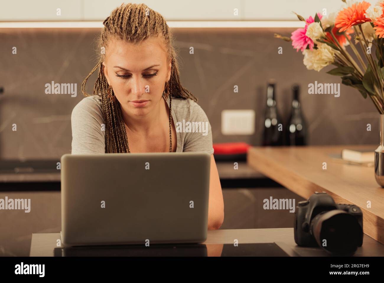 Junge Frau mit geflochtenem Haar, entweder eine professionelle oder Amateurfotografin, die eine DSLR und ein Notebook verwendet, experimentiert in der Küche, um sie zu meistern Stockfoto