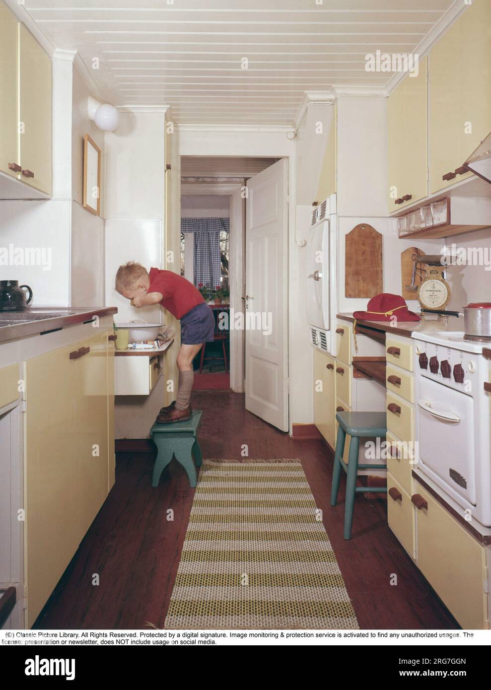 Küche der 50s-60s. Ein Junge, der auf einem Fußhocker steht und Hände und Gesicht wäscht. Die Küche ist typisch mit gelben Farben und sichtbaren Gegenständen aus der Zeit. Schweden 1950er 1960er Stockfoto