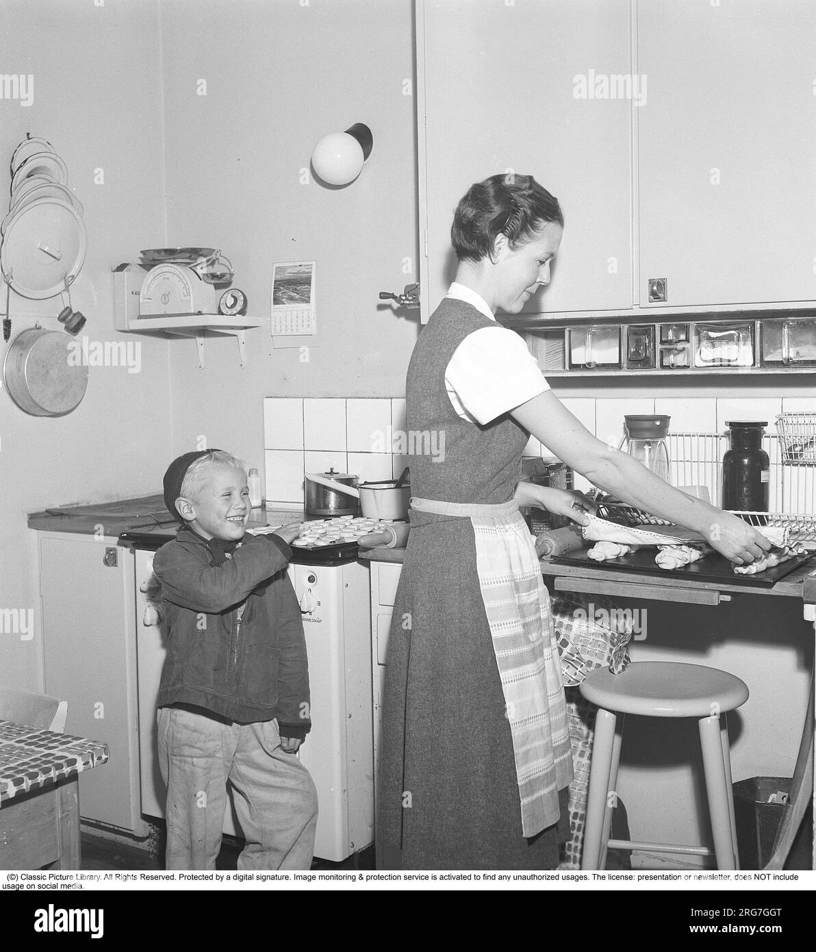 Backen in den 1950er. Eine Mutter mit ihrem Sohn in der Küche. Sie backt süßes Brot und Brötchen. Ein bereits gebackener Teller mit Kochis ist verlockend und der Junge streckt nach einem, wenn seine Mutter wegschaut. Schweden 1958 Kristoffersson Ref BY20-2 Stockfoto