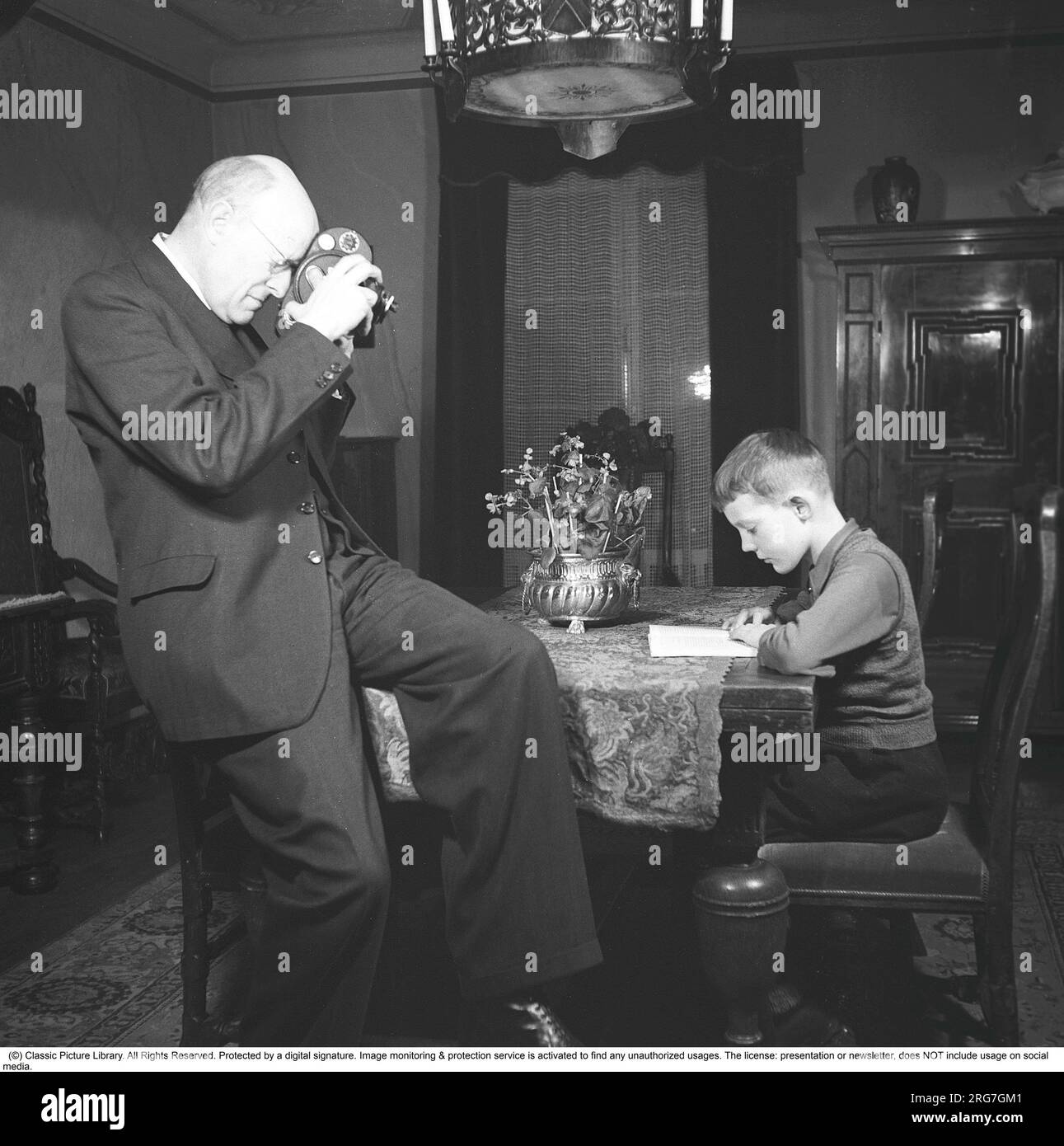 In den 1940er Jahren. Ein Mann filmt mit einer Amateurfilmkamera. Der Film wurde dann entwickelt und auf einem Filmprojektor auf einer klappbaren Leinwand zu Hause gezeigt. Der Film hatte keinen Ton. Die Filmkameras waren nicht batteriebetrieben und man musste sie wie eine Uhr aufziehen, damit sie funktionieren. Er konzentriert sich auf seinen Sohn, der an einem Tisch sitzt und liest. Schweden 1943. Kristoffersson Ref. B103-1 Stockfoto